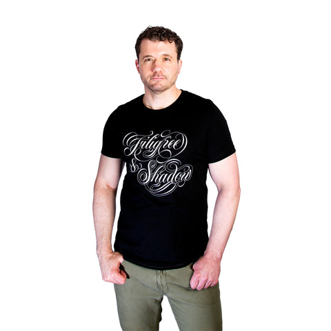 A man with short, curly hair wearing a black t-shirt with the Filigree & Shadow logo in white cursive lettering, standing against a white background.